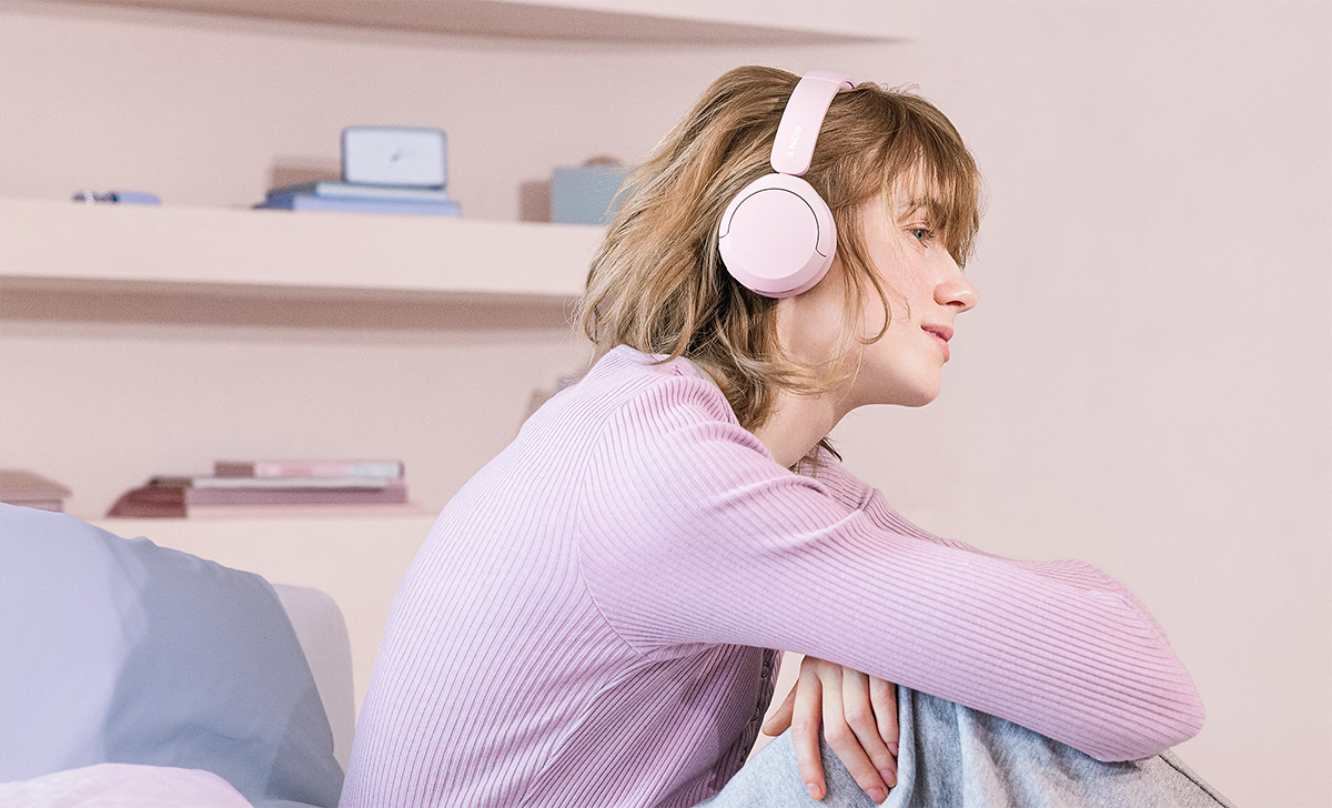 Mujer joven de costado sentada con los brazos apoyados en las rodillas flectadas usando los aud&iacute;fonos Sony WH-520 en color rosado