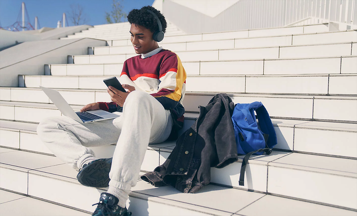 Hombre joven afroamericano sentado en unas escaleras, con notebook apoyado en su regazo y sosteniendo un smartphone en la mano izquierda, ambos emparejados con aud&iacute;fonos Sony WH-CH50