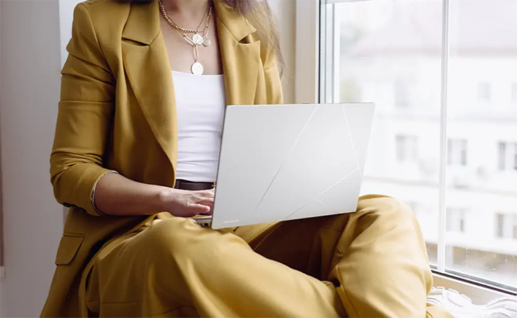 Mujer de traje sentada junto a ventana utilizando el dispositivo ASUS Zenbook 14 OLED apoyado en el regazo, destacando su gran capacidad de bater&iacute;a para una autonom&iacute;a que te acompa&ntilde;a durante toda tu jornada de trabajo