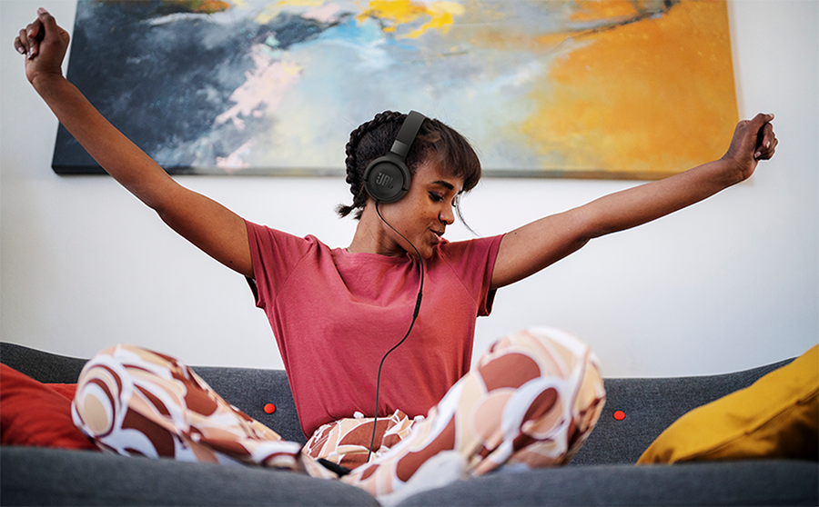 Mujer joven de color sentada sobre un sof&aacute; disfrutando de sus aud&iacute;fonos JBL Tune 520